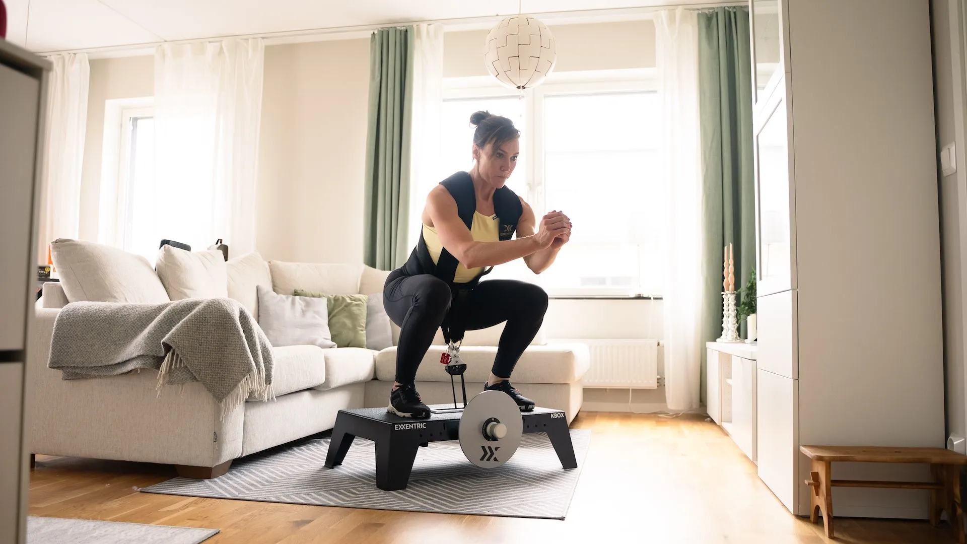 sara_squatting_on_kbox_active_with_harness_and_m_flywheel_in_the_living_room.webp